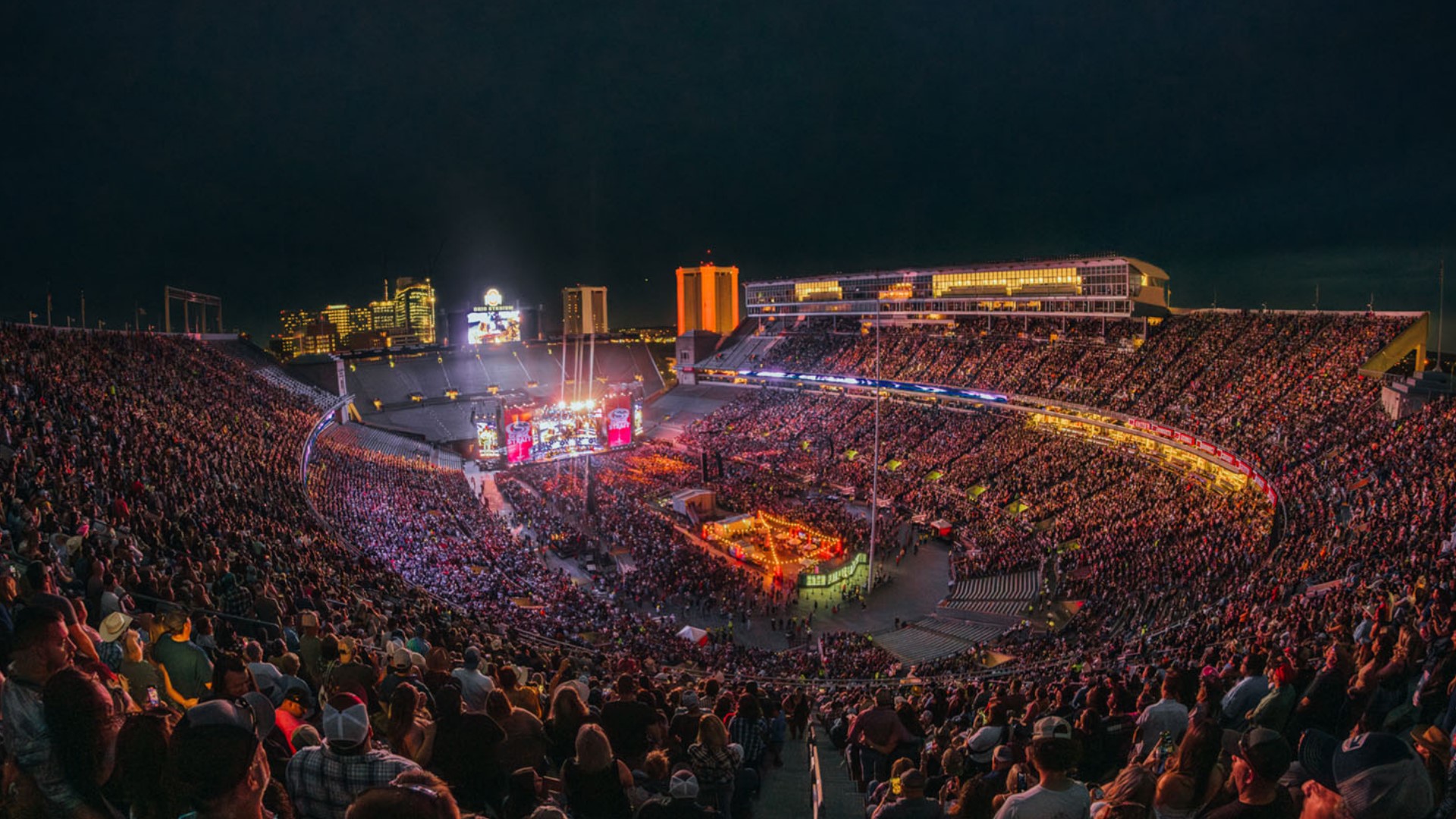 Packed stadium at night hosting a large concert with a lit stage on the field and a city skyline in the background.