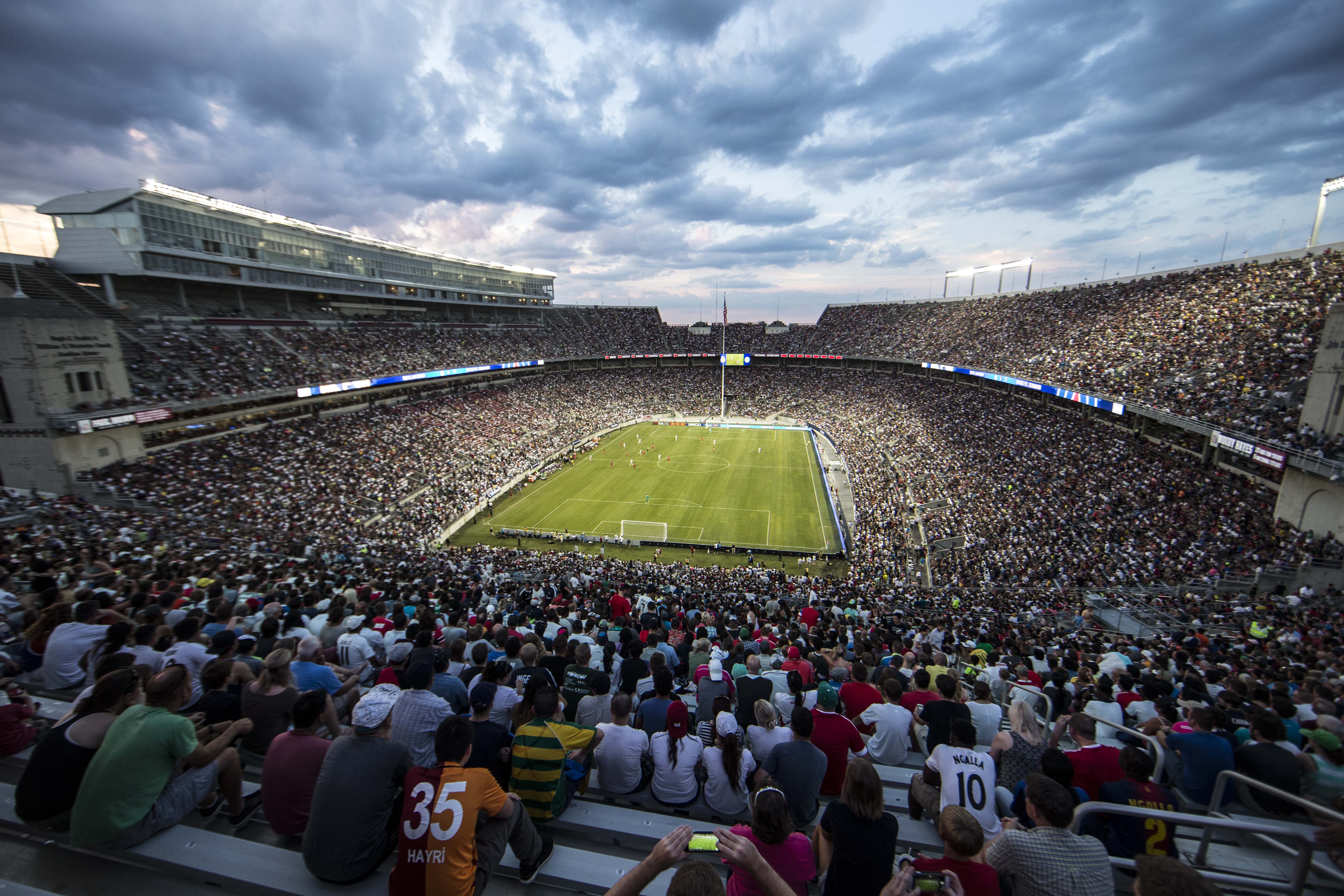 Aerial view of a packed stadium hosting a soccer match, with a green field and dramatic cloudy sky overhead.