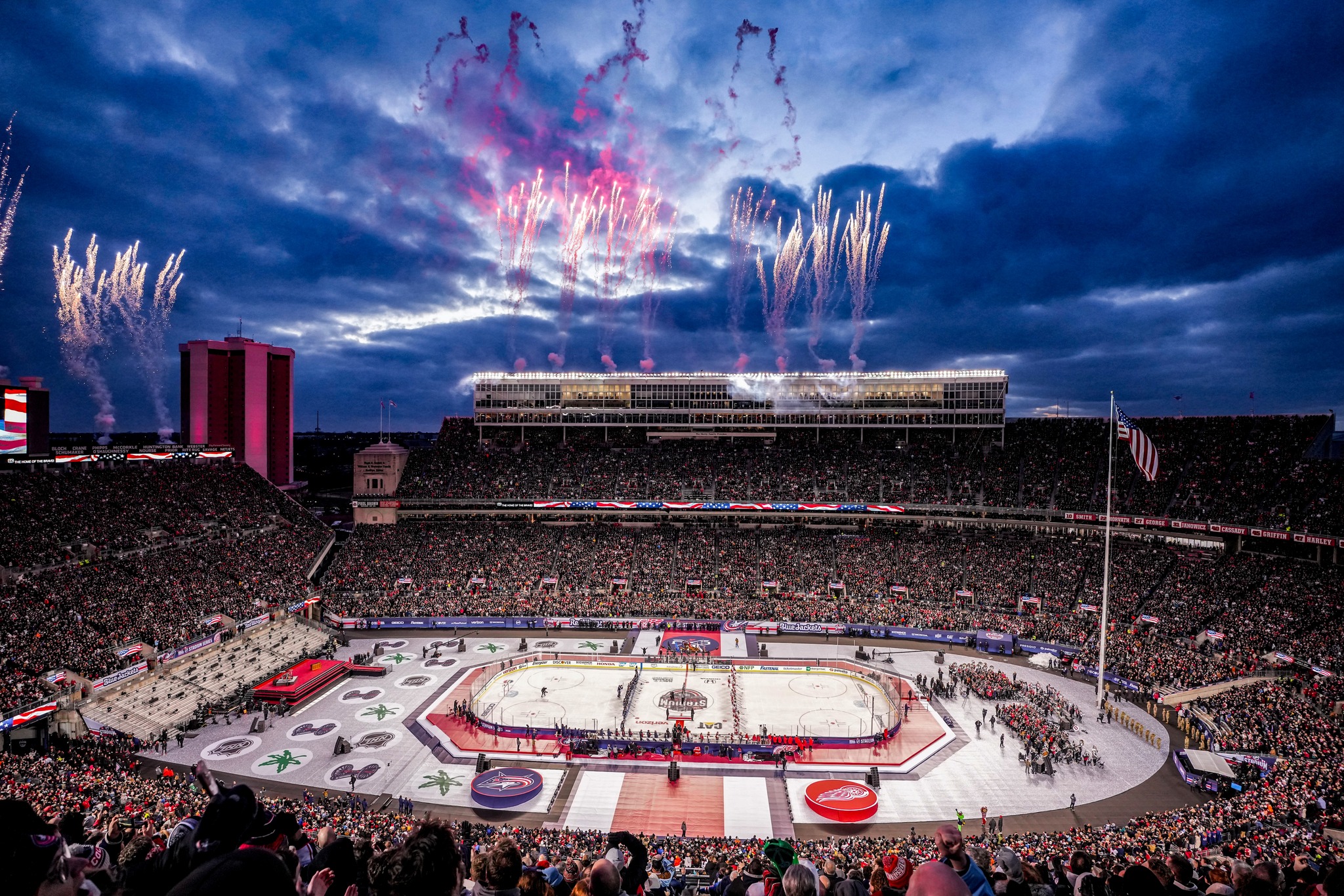 Packed outdoor stadium hosting an NHL hockey game at dusk, with fireworks bursting overhead.